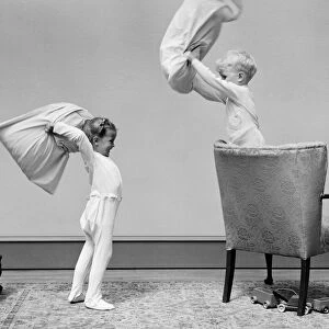 Boy and girl having a pillow fight, boy standing on chair swinging pillow, girl on floor