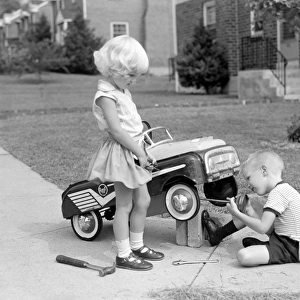 Children on suburban sidewalk, boy playing as mechanic, oiling toy pedal car