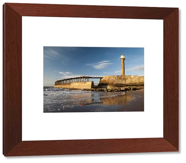 View of beach and pier with 19th century stone lighthouse in evening sunlight, East Pier, Whitby, North Yorkshire