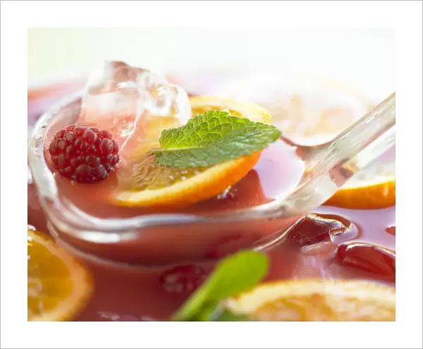 Plastic spoon scooping up punch from bowl, containing oranges, watermelon, raspberries, mint and ice cubes