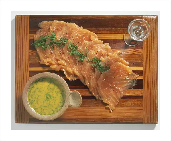 Bowl of sauce and thin slices of pink salmon seasoned with herb sprigs on wooden board, view from above