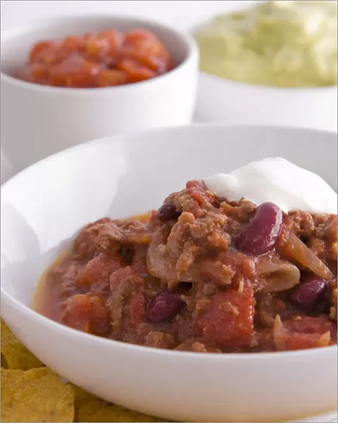 Bowl of chilli con carne with tortillas chips, soured cream and bowls of salsa and guacamole in background