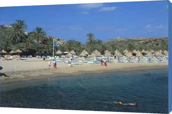 Greece, Crete, Vai Beach, sunloungers with thatched shades, calm waters and native palms, tourists dotted on sands
