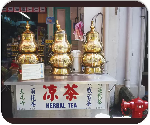 Singapore, Chinatown, Temple Street, decorative herbal tea dispensers, gold coloured metal decorated with Chinese script and dragons, lid loosely placed on central dispenser, standing on metal box, shop in background