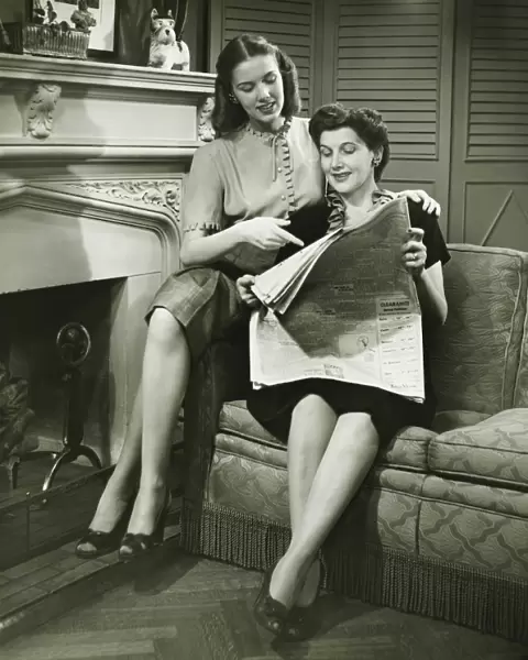 Two women sitting by fireplace, reading newspaper, (B&W)