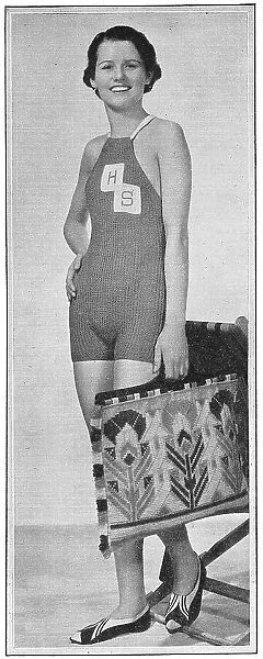 Model wearing a knitted bathing costume with HS monogram, carrying a beach bag worked in tapestry wools. Date: 1935