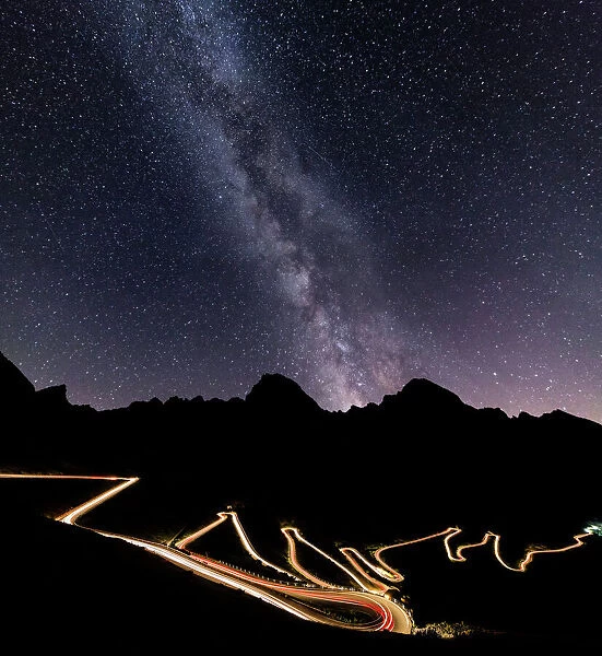 Panorama of the Milky Way and lights of car trace at Stelvio Pass, Valtellina, Lombardy