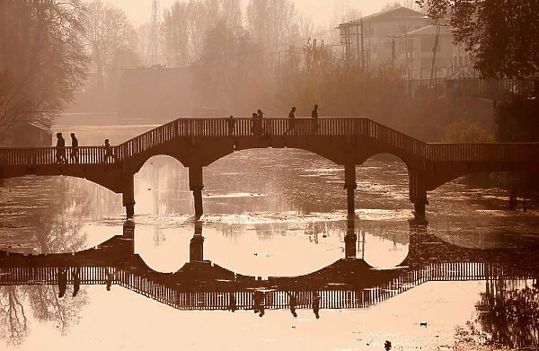 People walk on a wooden footbridge across a canal during an autumn day in Srinagar