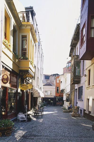 Asia, Turkey, Istanbul, cobbled Ortakoy side street lined with colourful shops and cafes, table and chairs outside, quiet scene