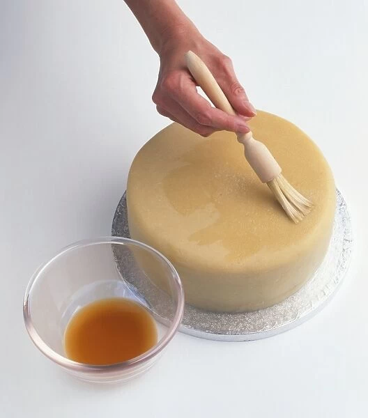 Person using basting brush to moisten cake topped with marzipan with brandy, close-up