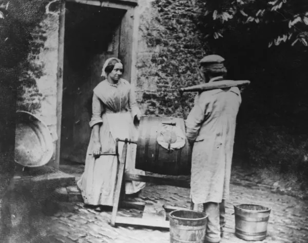 Butter Making. circa 1857: A woman churning butter talks to a milkman wearing