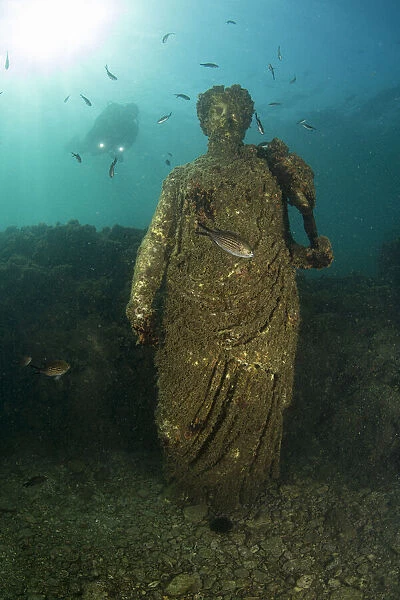 Scuba diver near ancient Roman Statue of Antonia minor, member of Julio-Claudian dynasty, daughter of Marcus Anthony and sister of emperor Augustus, in submerged Nymphaeum of Emperor Claudius. Marine Protected Area of Baia, Naples, Italy