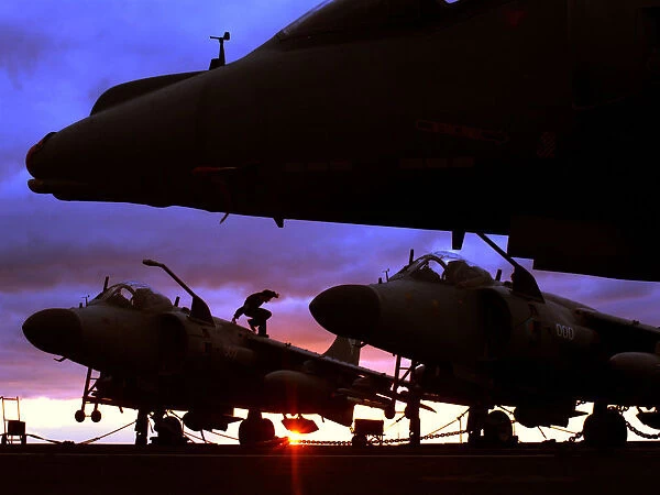 Harrier GR7s and Sea Harrier FA2s, are shown waiting on the deck of HMS Invincible at sunset