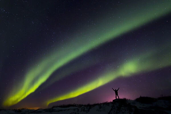 Colourful Aurora Borealis Over A Man With Arms Outstretched Silhouetted Against Light Pollution From Nearby Fort Greely; Alaska, United States Of America