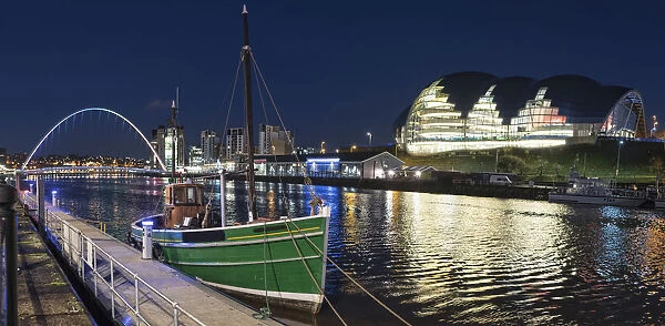 A Green Fishing Boat Moored Along The Edge Of A River At Nighttime With An Illuminated Sage Gateshead In The Background; Newcastle, Tyne And Wear, England