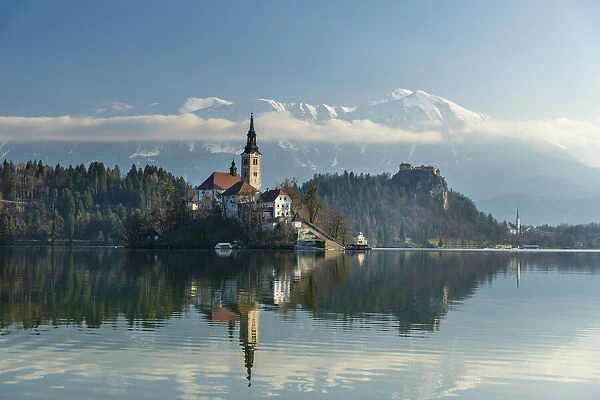 Looking across the calm waters of Lake Bled at dawn to the church on Lake Bled Island and Bled Castle behind; Bled, Slovenia