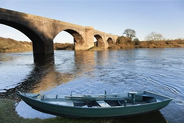 Northumberland, England; A Boat Moored On The Edge Of A River With A Bridge