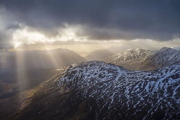 Sun breaking through cloud over snowy mountains near Glenfinnan; Scotland, United Kingdom