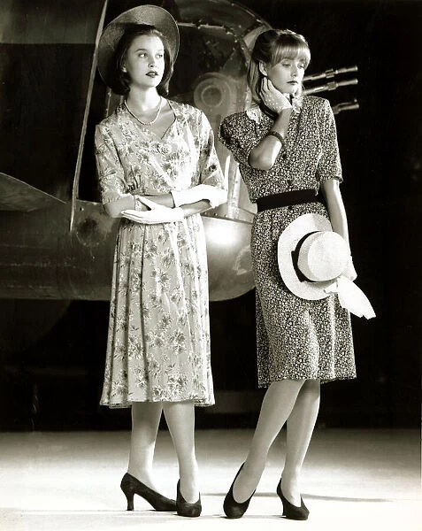 Two female models wearing 40s style evening dress with floral print holding a hat in one