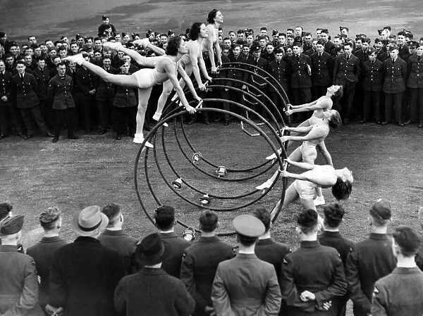 Women performing a gymnastic routine with large metal hoops, watched by military men