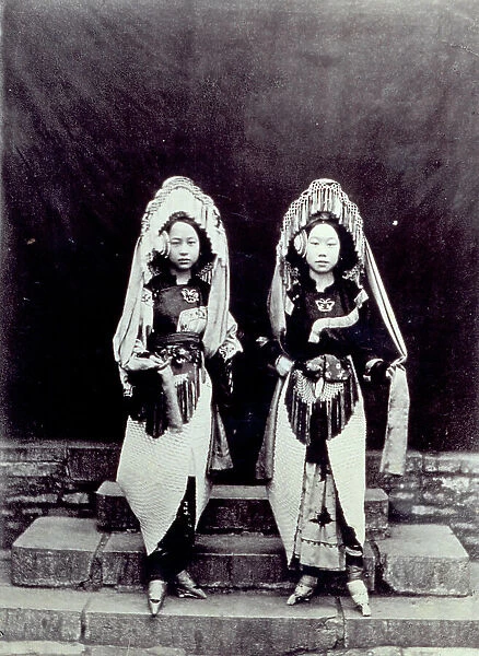 Full-length portrait of two small Chinese girls in traditional dress. They are showing their feet with the characteristic high-heeled pointed clogs