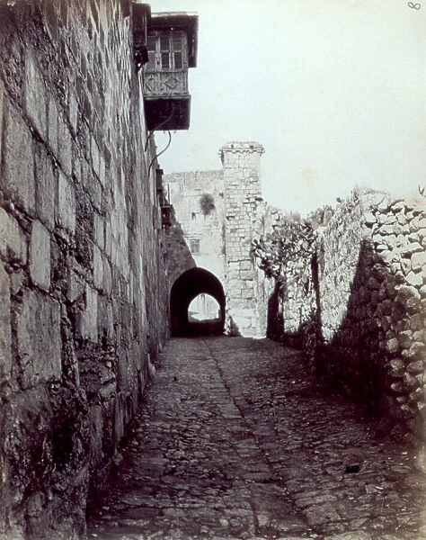 The ruins of the Antonia Fortress in Jerusalem. In the foreground, a narrow paved street, between a building and a tall wall with numerous cactus plants