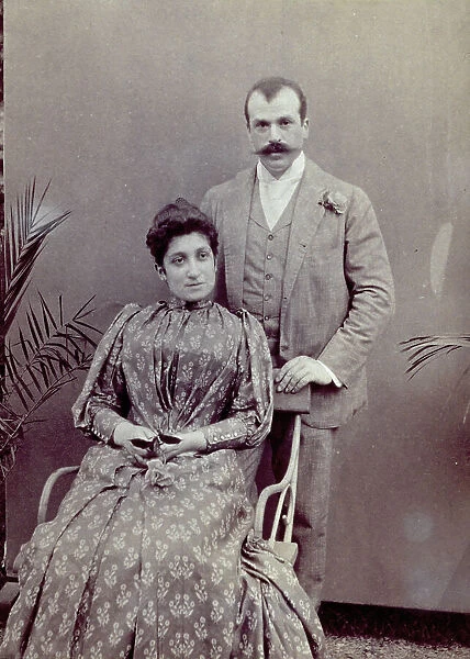 Studio portrait of a husband and wife. She is seated and elegantly dressed, holding a rose. He is standing behind her