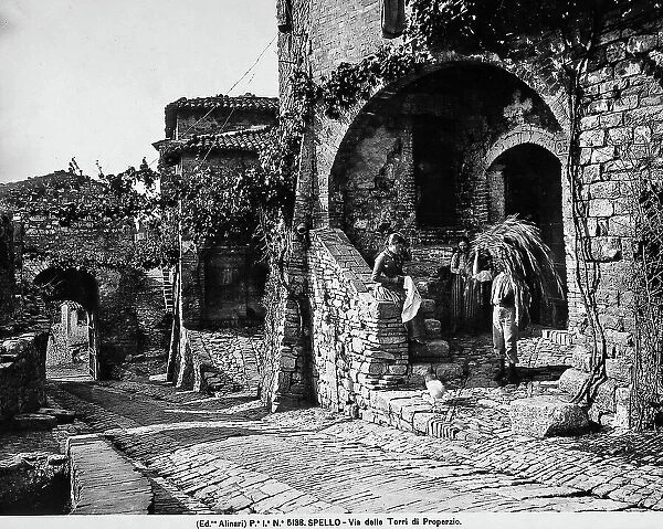 View of a part of the Via delle Torri di Properzio in Spello. In the foreground, a house is visible, in front of which are some farmers. One of them is holding sheaf of wheat