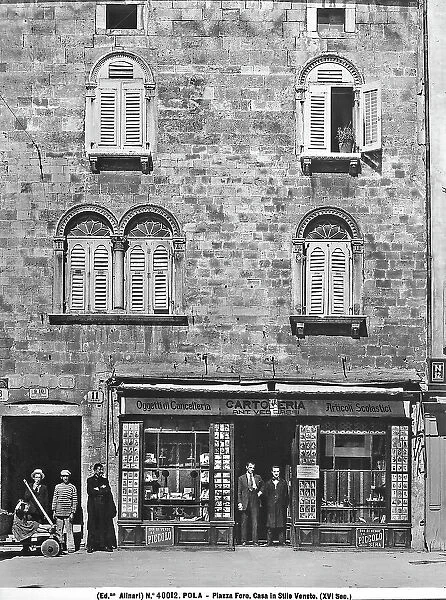 View of a facade in a Venetian style building from the XVI century, in Forum Square, Pula, photographed during the period of Italy's reign in Istria. There are men on the street in front of a stationer's shop