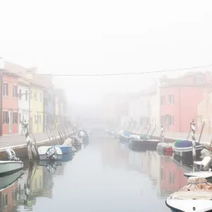 Colorful houses in Burano with canal and moored boats in the fog, Venice, Venetian lagoon