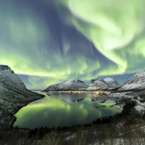 Northern lights and stars light up the snowy peaks reflected in the cold sea Bergsbotn