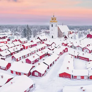 Pink arctic sunrise over the snowy red cottages and bell tower in the old Gammelstad Church Town, aerial view, Lulea, Sweden