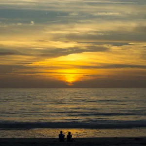 A couple sit on the sand watching the sun set on the Pacific Ocean in Cardiff