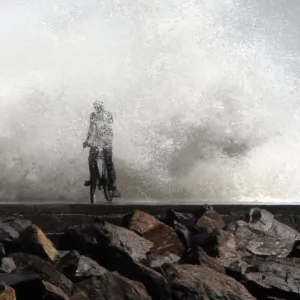 A man is drenched by a large wave during high tide as he cycles past at a fishing