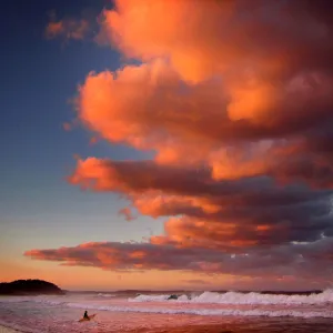 Surfer holding her board wades through the surf as clouds above are lit by the setting