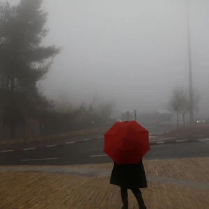 A woman holds an umbrella as she stands on the sidewalk during a foggy day in Jerusalem