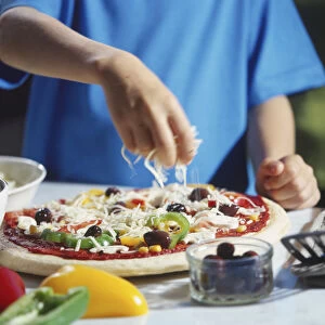 Cheese being sprinkled over pizza with vegetable toppings, surrounded by olives and sliced peppers in bowls, spatula