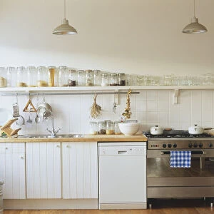 Kitchen with white wooden cupboards, pendant lights, long shelf stacked with glass jars containing dried foods, equipped with cooker and dishwasher, front view