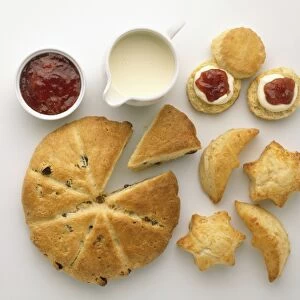 Large round fruit scone with slice removed, small star and crescent moon shaped cheese scones, traditional round scone with cream and jam toppings, dish of strawberry jam, small white jug of double cream, above view