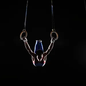 Teenage (16-17) male gymnast practicing on rings against black background