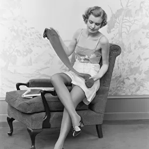 Woman wearing lingerie, sitting on arm of chair, holding up pair of silk stockings