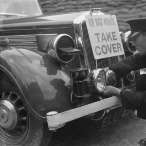 A policeman is fitting an air raid siren along with a warning sign to a police car