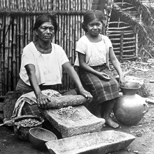 Historic image in black and white of a mother with young daughter rolling grist with rolling pin by hand to make tortillas, El Salvador circa 1920; El Salvador