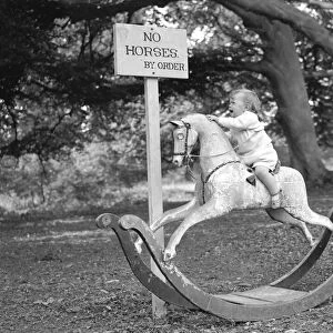 A baby riding a rocking horse beside a sign which reads No Horses by order. Circa 1940