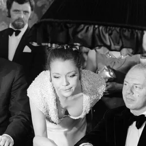 Diana Rigg in a low cut dress leans across the Black Jack table in a casino showing off