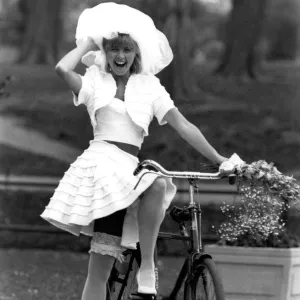 Rubbery Model Jane Sturdy. Wears white outfit, stockings and frilly hat on bicycle