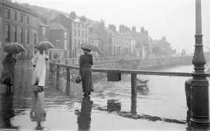 Prints of Pier Road, Whitby, North Yorkshire. People with raincoats and umbrellas walking beside the harbour in the rain. Photographed by Alfred Newton and Sons 1896-1920 Then and Now England - Historic Change Over Time: Rainy Day Scene, Whitby Harbour Print