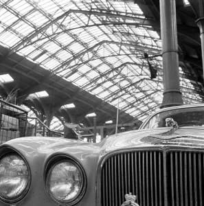 Prints of LIVERPOOL STREET STATION. London. Detail of the front of a Jaguar car parked within the station and the station roof in the background. Date range 1960-1972. John Gay Transport Prints: Liverpool Street Station Print, 1960-1972