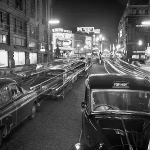 Prints of PICCADILLY, London. Stationary traffic along a busy Piccadilly at night, showing a London taxi cab for hire, and the illuminated signs of Piccadilly Circus and Lillywhites in the background Transport Prints: Nighttime Piccadilly Print, London