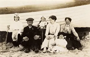 <b class=qtipBold><figcaption>Extended family group at the seaside</figcaption></b><br>Extended family group at the seaside - photographed with their backs to a fishing boat (perhaps to act as a windbreak?!) - note the 'News of the World' bucket held by the middle child - perhaps an early example of a marketing 'give-away' by the newspaper? Date: circa 1910s<br><em>© Grenville Collins Postcard Collection / Tom Gillmor / Mary Evans</em> Extended family group at the seaside